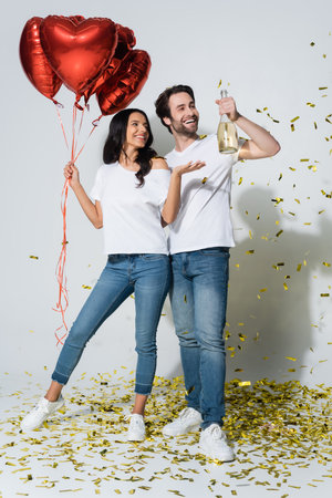 happy woman with heart-shaped balloons pointing at champagne bottle in hand of boyfriend on greyの写真素材