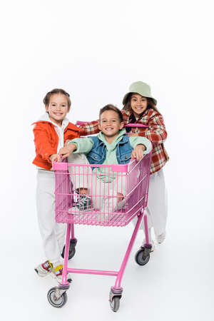 happy boy in shopping cart near joyful girls isolated on whiteの写真素材