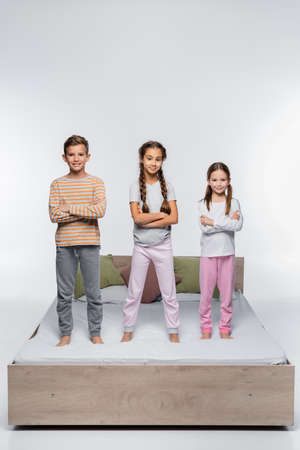 cheerful kids standing with crossed arms on bed and smiling on whiteの写真素材