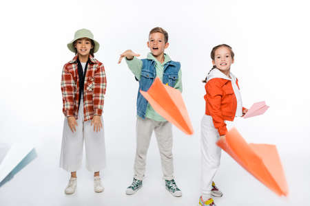 full length of happy preteen kids throwing paper planes in air on white