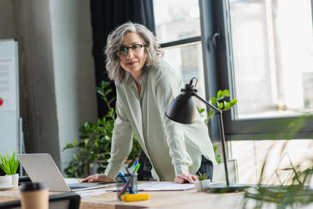 Grey haired manager looking at camera near laptop and documents on working table in officeの写真素材