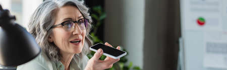 Grey haired businesswoman in eyeglasses recording voice message on smartphone near blurred lamp in...