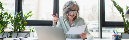 Mature businesswoman holding paper during video call on laptop, banner