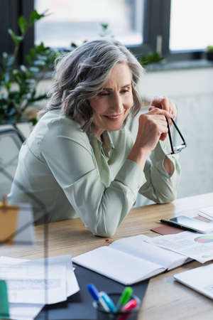 Cheerful businesswoman holding eyeglasses near documents and gadgets on table in officeの写真素材