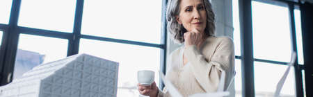 Thoughtful businesswoman holding cup near model of building in office, banner