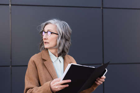 Mature businesswoman in coat holding paper folder and smartphone while looking away on urban streetの写真素材