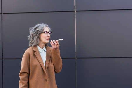 Mature businesswoman in coat recording voice message on smartphone outdoors