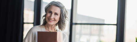Middle aged businesswoman holding paper folder and smiling at camera in office, banner
