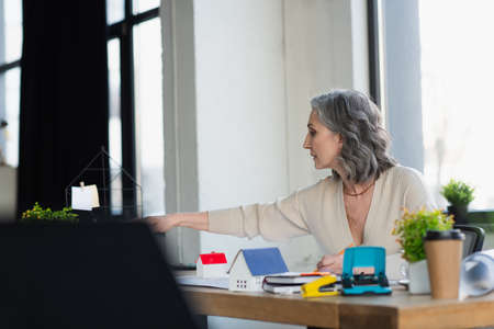 Side view of businesswoman working near models of houses and blueprints in office