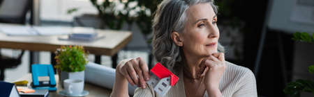 Pensive businesswoman holding model of house in office, banner