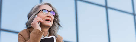 Grey haired businesswoman in eyeglasses talking on mobile phone and holding paper folder outdoors,...