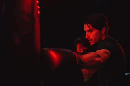Sportsman in boxing gloves training with punching bag on black background