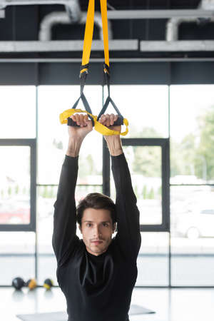 Brunette sportsman training with suspension straps and looking at camera in gym
