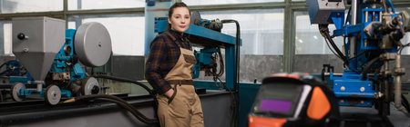 Young welder looking at camera near welding mask and machines in factory, banner
