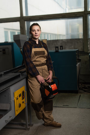 Young welder holding mask near welding machine in factoryの写真素材