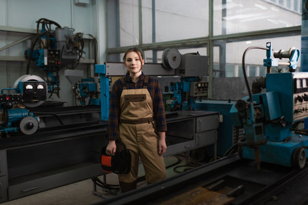 Brunette welder holding protective helmet near welding machines in factoryの写真素材