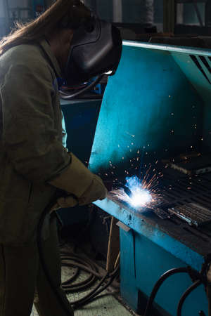 Young welder in uniform and protective mask working with welding torch in factory