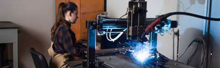 Side view of welder in overalls sitting near keyboard and welding machine in factory, banner