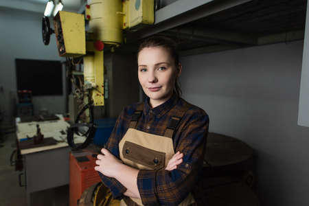 Pretty welder in overalls looking at camera near blurred welding machine in factory