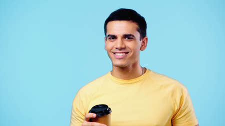 cheerful young man in yellow t-shirt holding paper cup isolated on blue