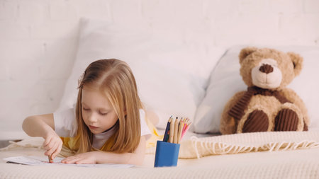 Kid drawing with color pencils near blurred teddy bear on bedの写真素材