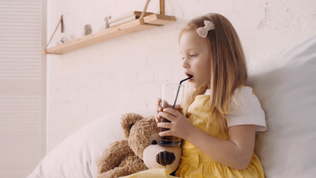 Kid holding glass of juice near teddy bear on bed