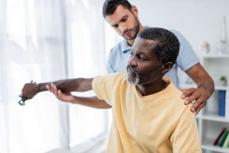 mature african american man exercising with dumbbell near blurred physical therapistの写真素材