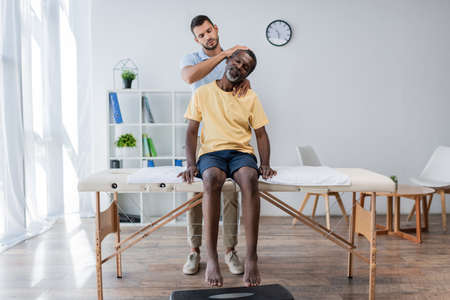 doctor examining mature african american man sitting on massage table in rehabilitation centerの写真素材