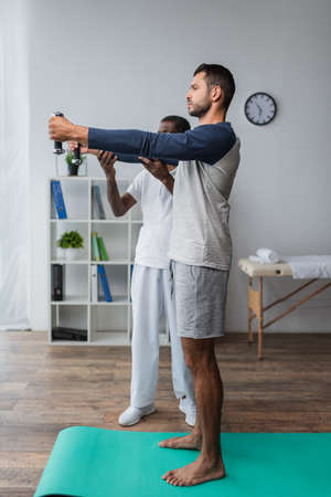 african american rehabilitologist helping young barefoot man working out with dumbbellsの写真素材