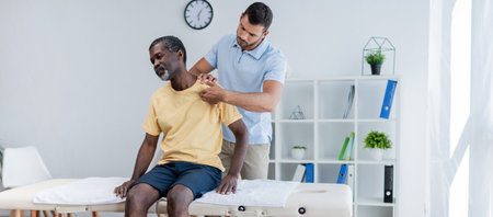 middle aged african american man sitting on massage table while rehabilitologist examining his shoulder, bannerの写真素材