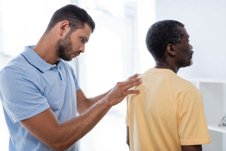 young rehabilitologist examining shoulder and back of mature african american man