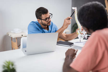 physiotherapist in eyeglasses pointing at spine model near blurred african american patientの写真素材