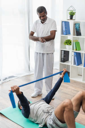 african american rehabilitologist looking at young man training with rubber band while lying on fitness matの写真素材