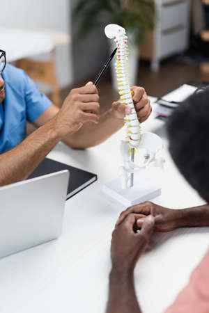 partial view of doctor pointing at spine model near blurred african american man in rehabilitation...