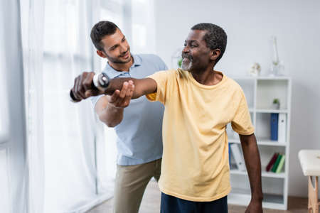 young trainer assisting happy african american man training with dumbbell in rehabilitation centerの写真素材