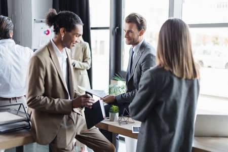 smiling african american businessman holding book while standing near business partners in office