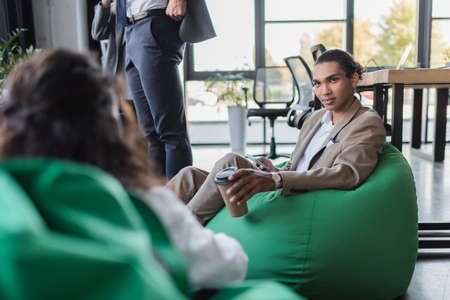 african american businessman with coffee to go sitting in bag chair near blurred colleagueの写真素材