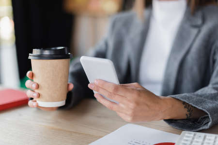 cropped view of businesswoman sitting at workplace with takeaway drink and cellphone