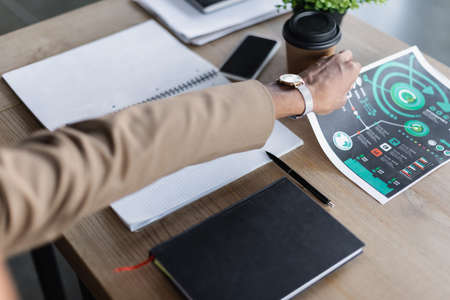 cropped view of african american businessman taking infographics near notebooks, coffee to go and...
