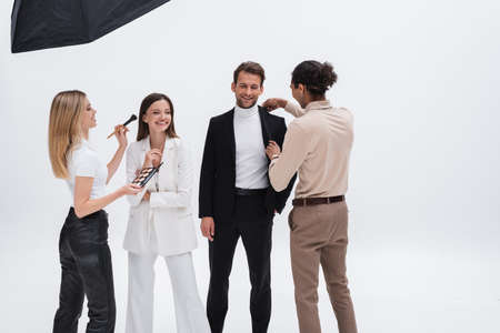 stylish models smiling near makeup artist and african american stylist preparing them to photo...