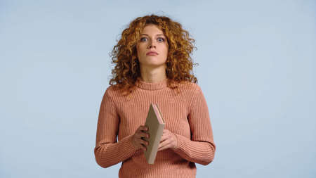 shocked young woman in pink sweater holding book while looking at camera isolated on blue