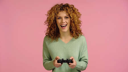 KYIV, UKRAINE - DECEMBER 22, 2021: excited redhead woman gaming isolated on pink