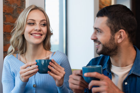 happy woman looking at blurred man while drinking coffee in restaurant