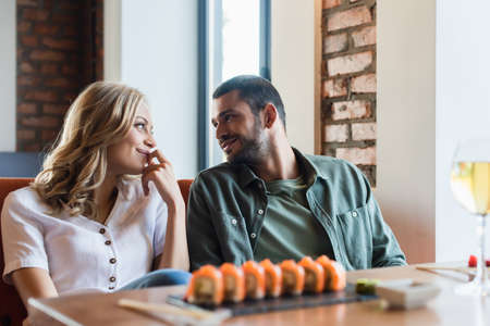 joyful couple smiling at each other near blurred sushi rolls and glass of white wineの写真素材