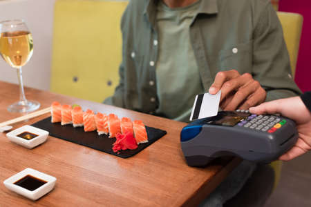 cropped view of blurred man paying through credit card reader near sushi rolls and wine glassの写真素材