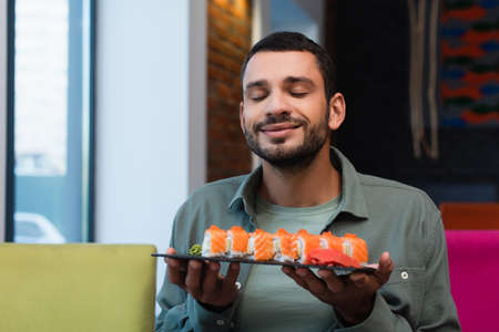 pleased man with closed eyes holding plate with tasty sushi rolls in restaurantの写真素材