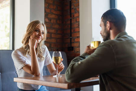 joyful woman looking at blurred man while drinking white wine in restaurant