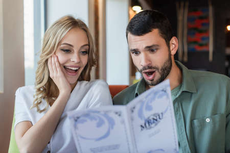 shocked woman touching face while reading restaurant menu near surprised boyfriendの写真素材