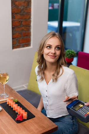 smiling woman with credit card paying through terminal for sushi rolls and glass of wine