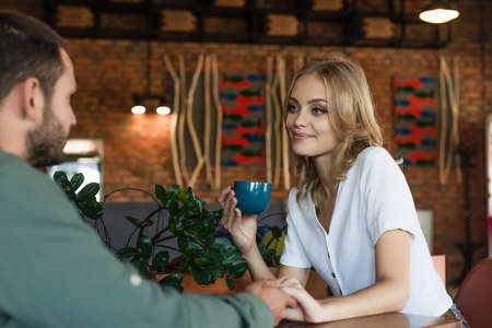 pleased blonde woman with coffee cup holding hands with blurred boyfriend in cafeの写真素材